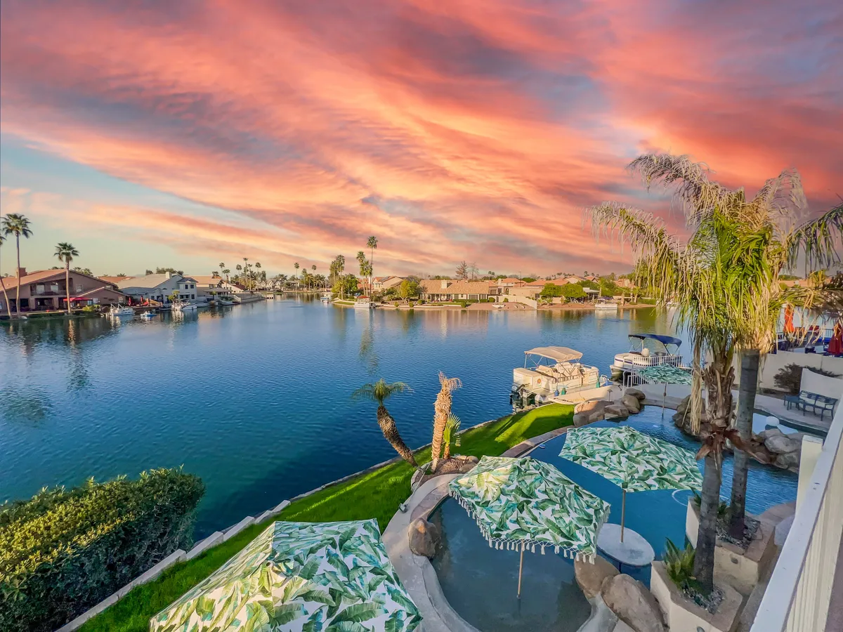 A peaceful lakeside scene featuring calm water, palm trees, decorative umbrellas, and outdoor seating, all illuminated by a colorful sky. The ambiance suggests relaxing afternoons by the lake, memorable gatherings, and an emphasis on community and leisure.