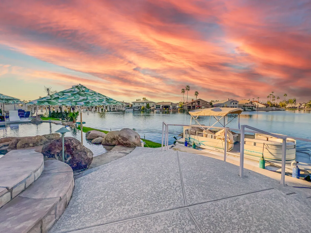 A tranquil waterfront scene at sunset with a boating area, landscaped features, and rich, colorful skies reflecting on the calm water. The setting invites peaceful evenings, boating, and social gatherings along the shore.