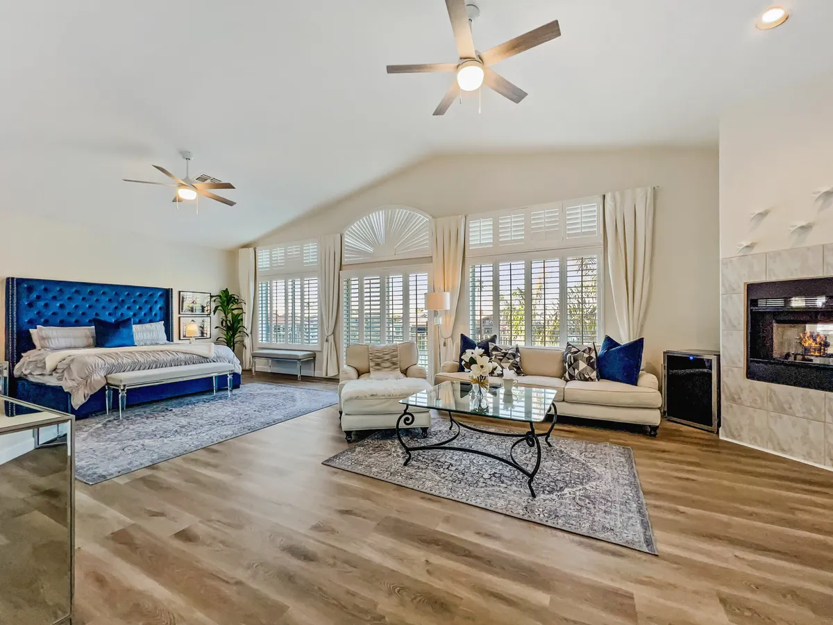 A spacious and comfortable king bedroom featuring a blue tufted bed, glass coffee table, large windows with plantation shutters, and a fireplace. The room is filled with natural light and exudes an air of elegance and relaxation, suggesting a primary suite with both comfort and luxury.