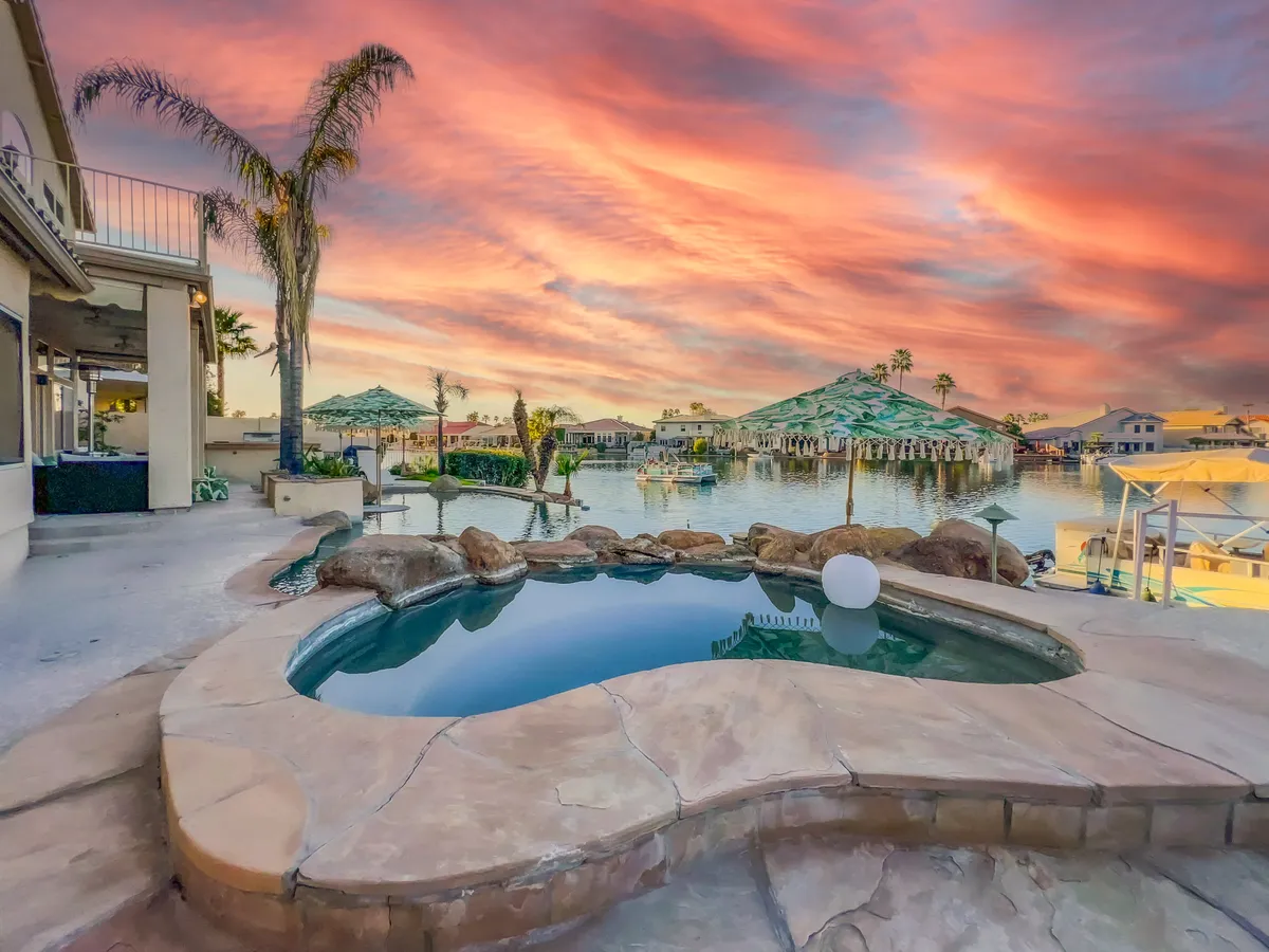 The photo features a bubbling hot tub in the foreground overlooking the water, framed by palm trees and a radiant sunset sky. Surrounding homes and lush landscaping add to the tranquil and exclusive atmosphere.