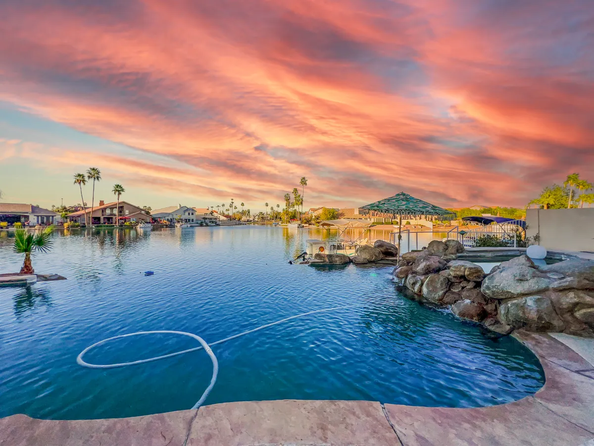 A stunning waterfront scene at sunset, with calm water reflecting a vibrant sky, palm trees bordering the shore, rock formations adding texture, and elegant houses in the background. The tranquil setting hints at opportunities for swimming, boating, and relaxing by the water's edge.