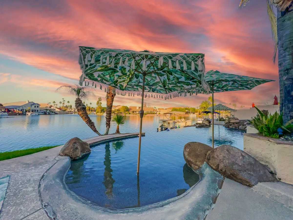 This image captures a stunning outdoor pool surrounded by natural stones, palm trees, and decorative umbrellas. The tranquil water reflects the colorful sunset sky, creating a resort-like ambiance ideal for unwinding or entertaining guests. The scene suggests a luxurious space for swimming, relaxing, or socializing by the poolside while enjoying breathtaking sunset views.