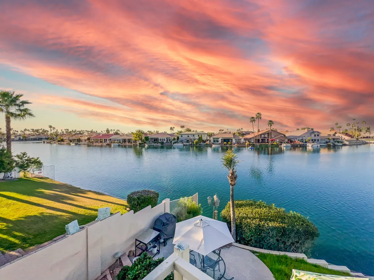 A serene view across calm waters at sunset, with waterfront properties and stylish patio furniture in the foreground. The colorful sky and placid water create a tranquil backdrop for outdoor dining, evening conversations, or special gatherings.