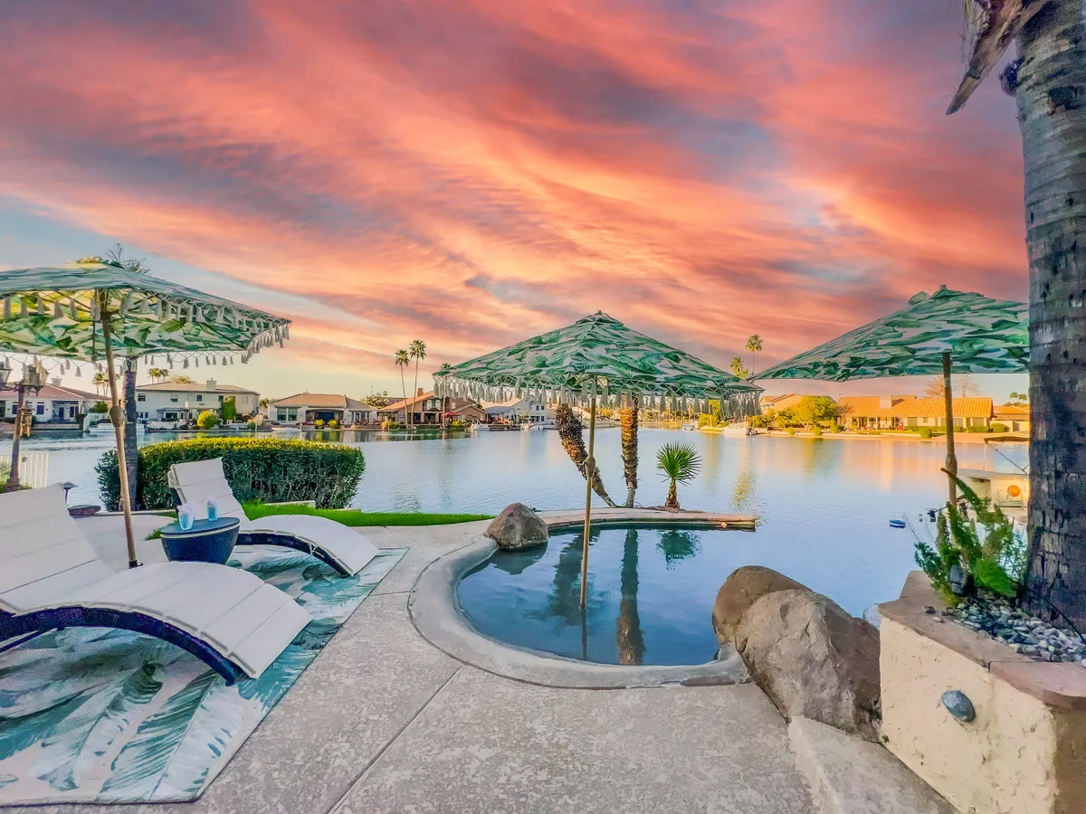 A serene exterior view featuring palm trees silhouetted against a vibrant sunset sky, a sparkling heated pool surrounded by lounge chairs and beach umbrellas, all set beside a calm waterfront. The inviting set-up suggests relaxation, sunbathing, and socializing by the water's edge.