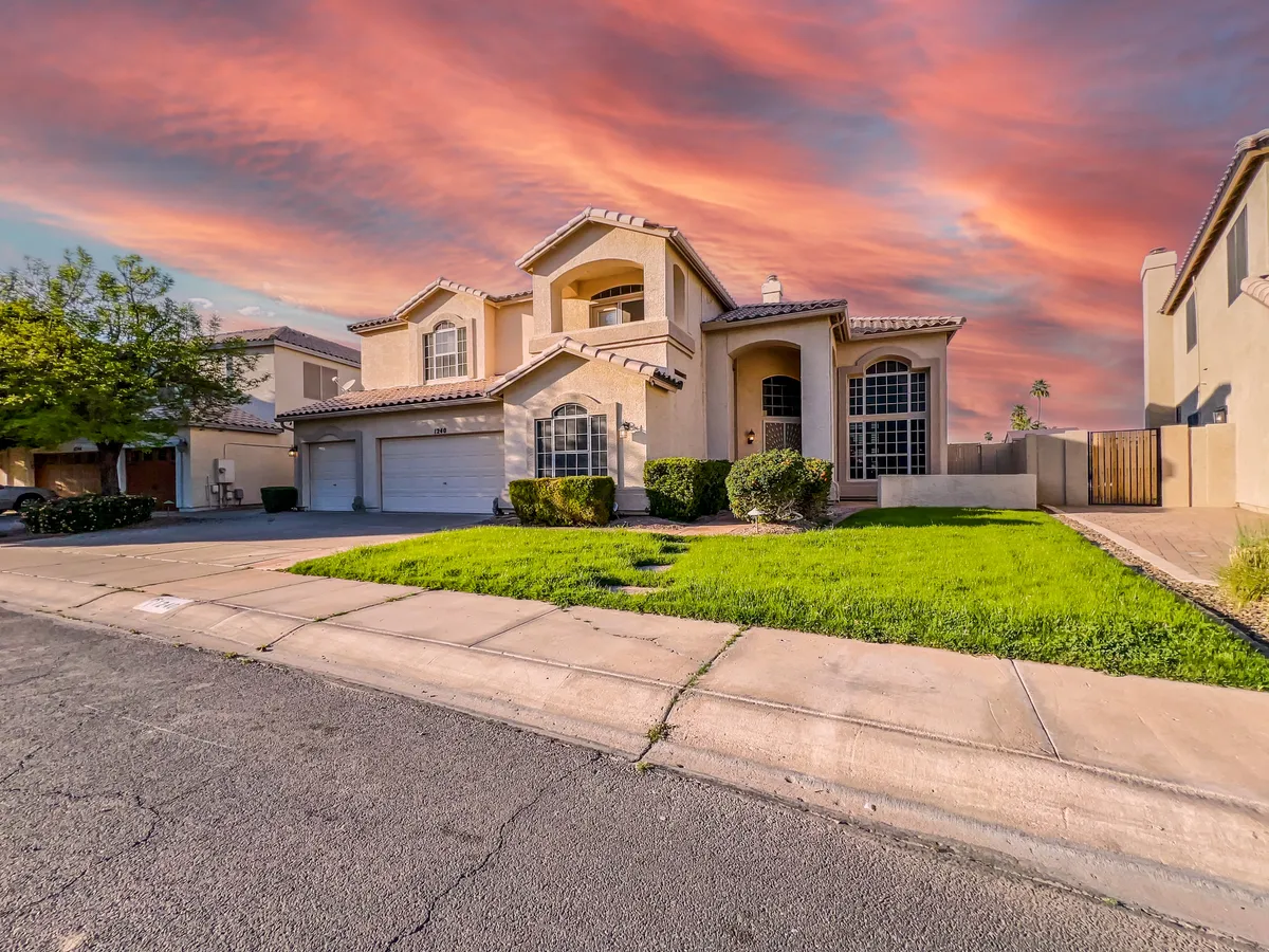 A welcoming front exterior view of the property featuring a large front yard, arched windows, a spacious two-car garage, and textured roof tiles, all basking in bright Arizona sunlight. The overall mood is inviting and homey, hinting at a comfortable stay for extended relaxation.