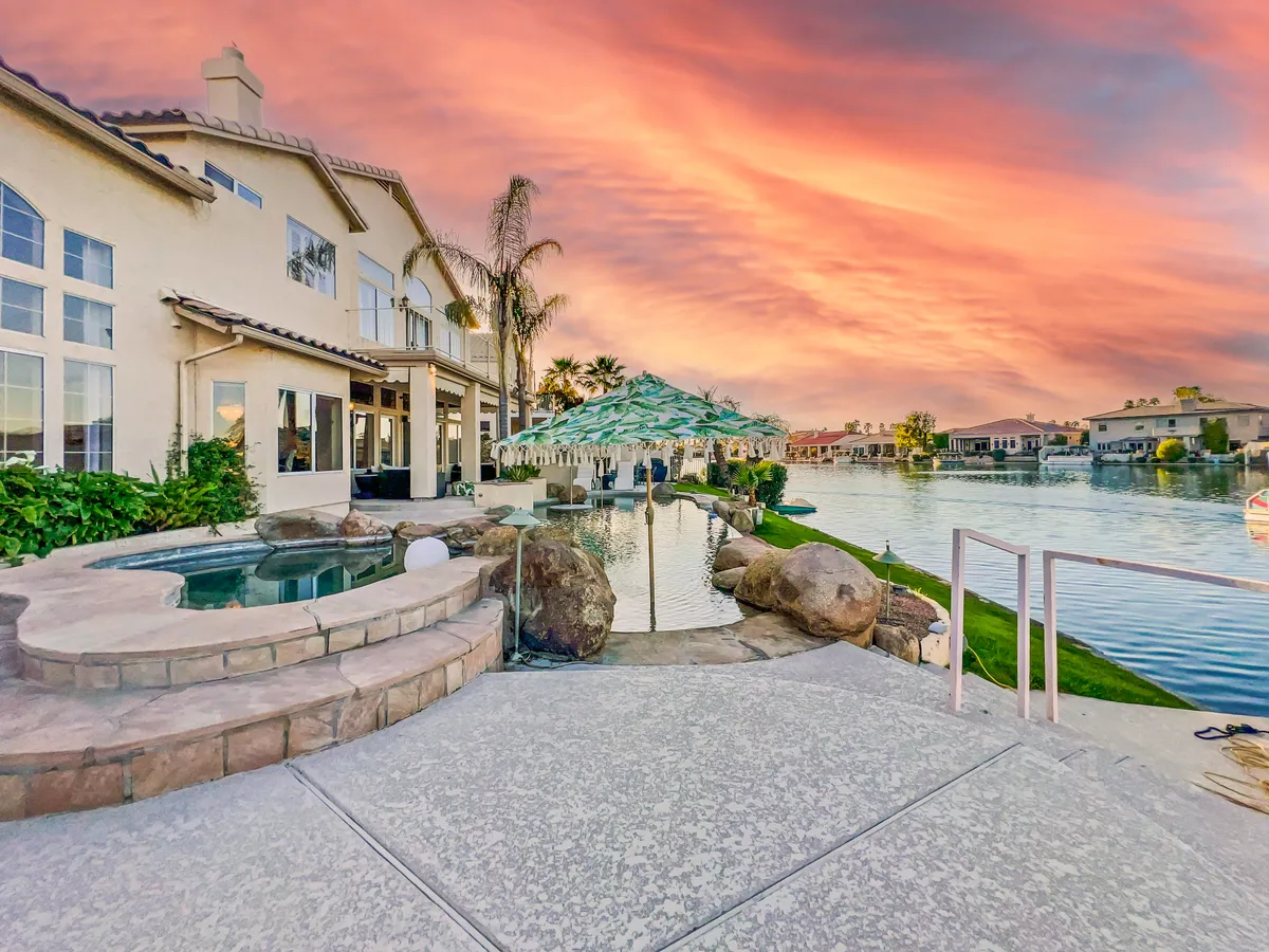 This exterior shot captures a stunning poolside scene at sunset, complete with the waterfront just beyond, a beautifully maintained patio area, and umbrella-covered seating. The tranquil water and open sky evoke a true sense of resort-style relaxation and leisure.